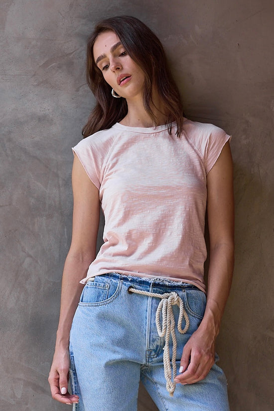 Woman wearing a pink t-shirt and light blue cut off jean shorts against a textured wall.