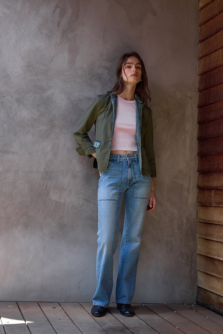 Woman wearing a green jacket with pink tank and blue jeans standing against a textured wall.