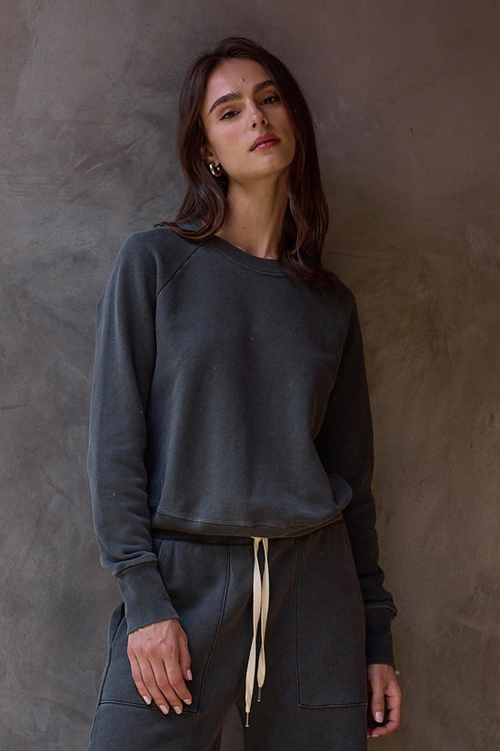 Woman wearing a dark gray sweatshirt and pants against a textured wall.