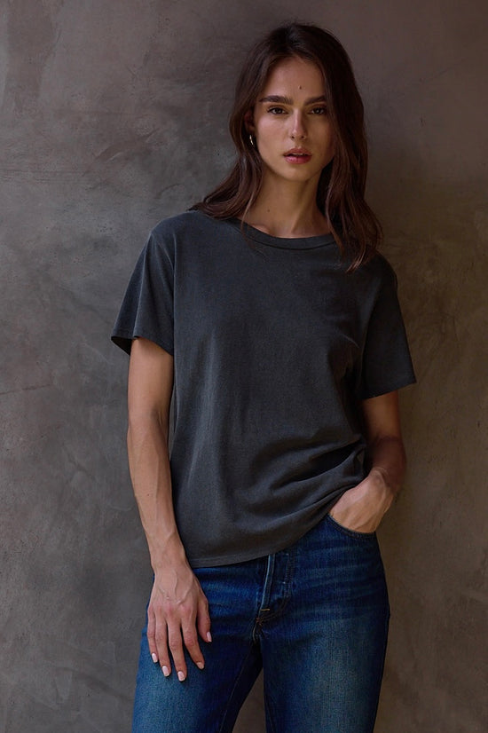 Woman wearing a dark gray t-shirt and blue jeans against a textured brown wall.
