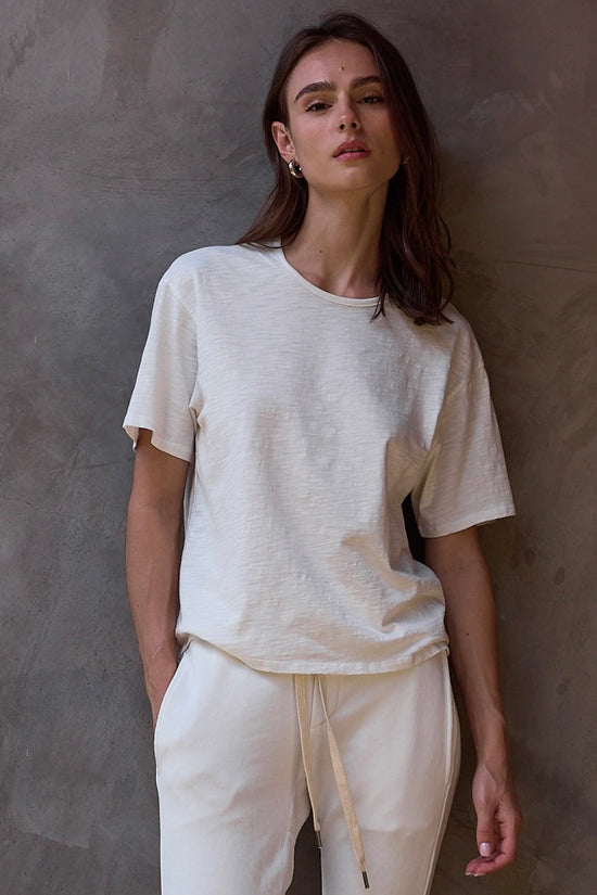 Woman wearing a soft white t-shirt and sweatpants against a textured wall.