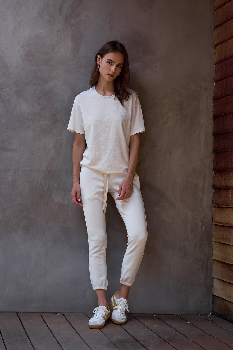 Woman wearing a soft white t-shirt and sweatpants standing against a textured wall.