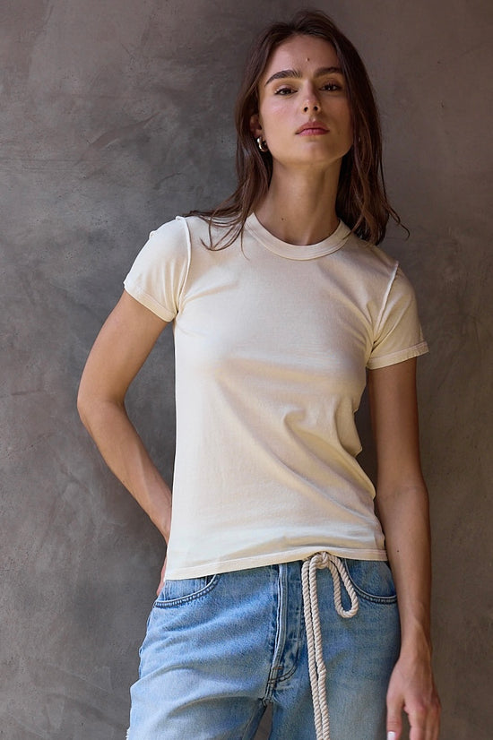 Woman wearing a pale yellow t-shirt and blue jeans against a textured wall.