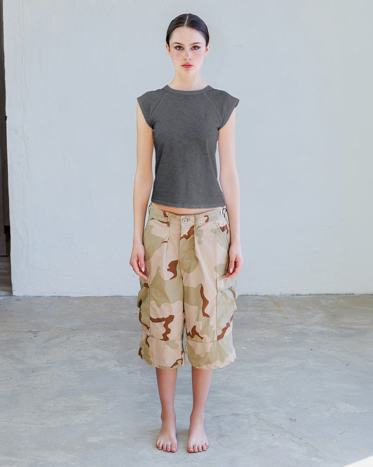 Woman wearing a gray top and camouflage shorts standing against a plain background